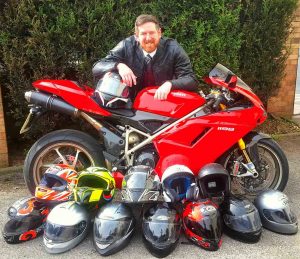 Funeral director leaning on red motor cycle. In front there are 12 helmets.