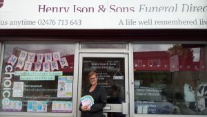 Picture of women standing in front of Henry Ison & Sons shop window. She is holding up a leaflet advertising Dying Matters.