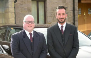 Picture of two gentlemen in suit and tie standing in front of a black car.