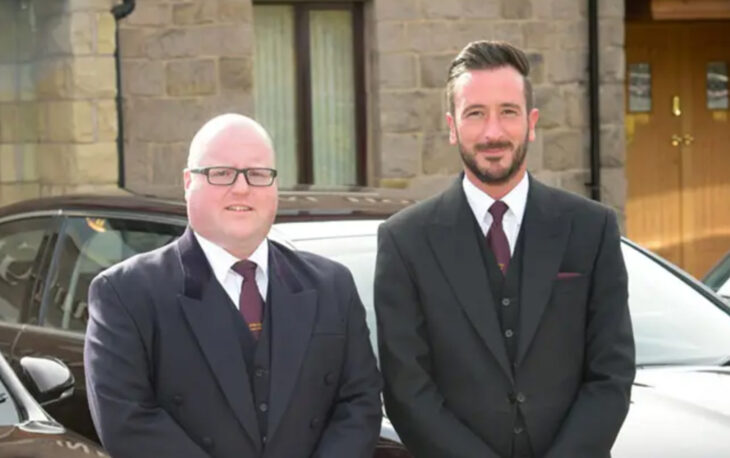 Picture of two gentlemen in suit and tie standing in front of a black car.