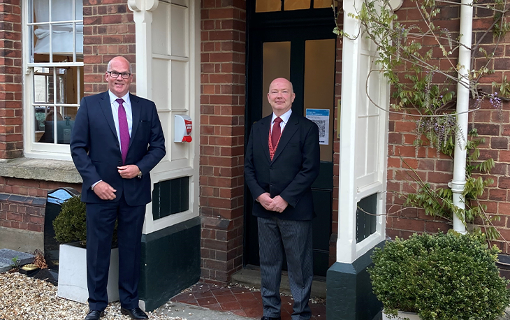 Picture 2 men in black suits and red ties in front of black wooden door.