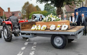 Picture of a tractor carrying a coffin to the burial with letters spelling dad and Sid in white and yellow flowers.