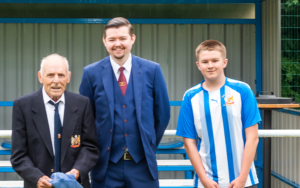 3 people are pictured in front of football stand. Two are in suit and tie and one is in the Wythenshawe FC kit.