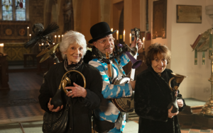 3 people standing in a triangle formation in front of church altar, two are holding honk horns and one is pretending to play the banjo.