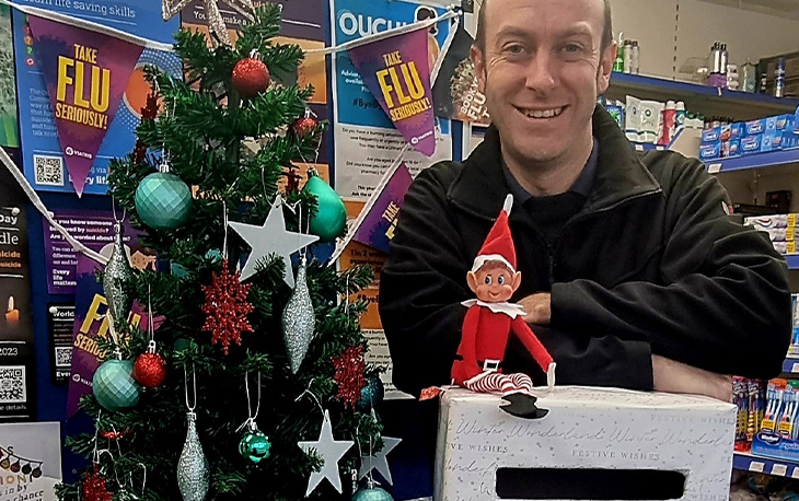 Man is pictured next to Christmas tree with small red Christmas elf in front of him.