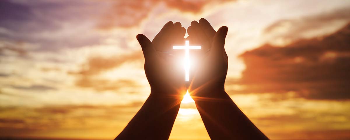 Hands holding up a glowing cross with a sunset in the background