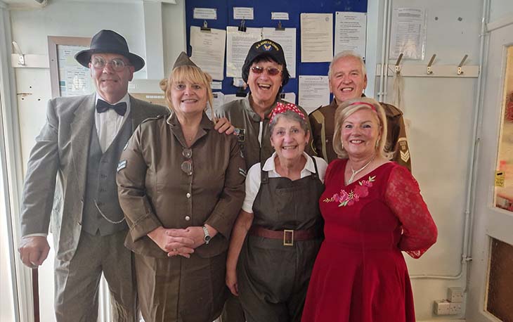 6 people gathered round dressed in olden day wear posing for a picture.
