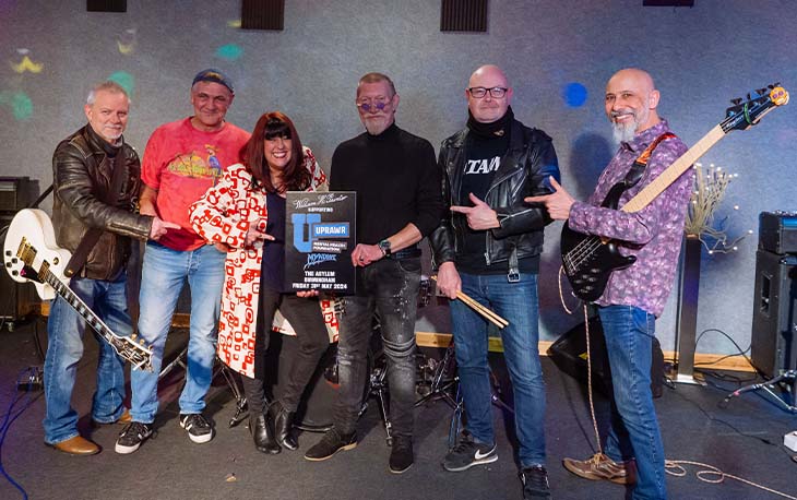 A band with 2 guitarists, 2 singers and a drummer holding up a sign for the UPRAWR Mental Health Foundation.